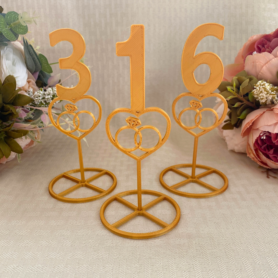 Gold table numbers that is 3D printed with a heart design that encloses a set of wedding rings